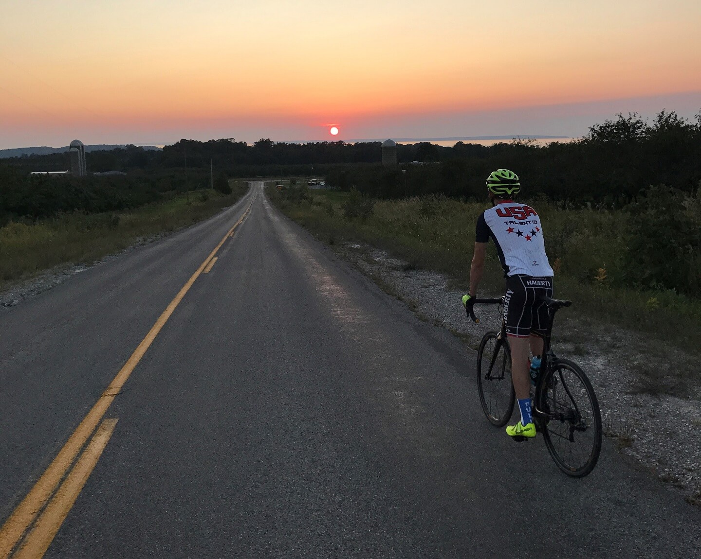 Young man on bicycle faces open road, sunset on horizon